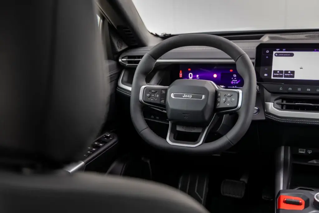 La tercera generación del Jeep Compass llega con una perspectiva global, fusionando el diseño icónico de la marca con un elevado nivel de equipamiento y una amplia dotación tecnológica pero, sobre todo, con una calidad de rodadura adaptada a los estándares europeos y, por qué no decirlo, con una puesta a punto perfecta para nuestras hoy maltrechas carreteras. Lo primero que llama la atención es que este Jeep Compass –que hemos probado en su versión eléctrica de lanzamiento– es claramente más grande que su predecesor y muestra un aspecto rotundo y cúbico que ha obligado a trabajar cada centímetro cuadrado de la carrocería para lograr un coeficiente aerodinámico de 0,29, meritorio para unos volúmenes tan poliédricos. El Jeep Compass se desmarca de sus “mellizos” de Stellantis Con 4,55 metros de longitud, Jeep lo define como SUV del segmento C, aunque sus dimensiones lo acercan claramente al segmento superior. Basado en la plataforma modular STLA Medium de Stellantis –compartida con los Peugeot 3008 y 5008, el Opel Grandland y el Citroën C5 Aircross–, el Jeep Compass se beneficia de un desarrollo de suspensiones específico orientado a lograr 20 centímetros de altura libre al suelo, con una reducción del 15 % en la aceleración vertical y un 20 % menos de balanceo de carrocería respecto a la generación anterior. El resultado sobre el asfalto es el de un buen SUV: no especialmente ágil, como corresponde a su masa y a su centro de gravedad elevado, pero sí notablemente mejor que el anterior Jeep Compass. Se inclina moderadamente en los apoyos fuertes y en las frenadas enérgicas, pero sin el efecto rebote de las suspensiones que caracterizaba a la generación precedente, poniendo en valor el trabajo de unos amortiguadores perfectamente capaces de cumplir con su trabajo. Las suspensiones están, así, bien adaptadas para recorrer caminos a buen ritmo: absorben con solvencia las irregularidades del terreno y muestran una parte final del recorrido más firme para evitar topes si se aborda una zona abrupta a más velocidad de la debida. En los caminos moderadamente rotos, el Compass pone en valor sus amplios recorridos de suspensión y la amabilidad con la que trata a sus ocupantes, y en autopista cumple con eficacia. En unos tiempos en los que nuestras carreteras acumulan socavones y baches por la falta de mantenimiento y el paso de las últimas borrascas, un vehículo como este Jeep Compass resulta más interesante de lo que parece. La dirección, además, ofrece un tacto suave, muy asistido, que minimizará el efecto “latigazo” ante un bache imprevisto, y el radio de giro es razonable para la batalla del vehículo. Al volante del Jeep Compass eléctrico, el motor de 213 CV ofrece la respuesta inmediata que cabe esperar de esta tecnología, pero con una progresión suave y lineal que casa bien con el carácter del vehículo. La frenada regenerativa está bien integrada, y el modo One Pedal –que detiene el vehículo por completo– es muy práctico, aunque resulta algo llamativo que se active mediante un botón dedicado en lugar de mediante las levas, como los otros tres niveles de regeneración. En cuanto a la autonomía homologada de 500 km WLTP, conviene aplicar el habitual factor corrector del 0,7 para hacerse una idea realista del alcance en uso cotidiano. Salto de calidad a bordo del nuevo Jeep Compass Por calidad de acabados y espacio, el habitáculo no tiene nada que ver con lo visto hasta ahora en el Jeep Compass. El puesto de conducción es cómodo, con una postura elevada muy Jeep, y las plazas traseras son amplias y adecuadas para el uso diario. El maletero ofrece 550 litros –45 más que en el anterior Jeep Compass–, ampliables con el gran hueco bajo el piso desmontable, aunque no hay maletero delantero. Un detalle relevante: el selector del cambio es un mando giratorio físico e intuitivo, en lugar del habitual conmutador lineal del resto de modelos de Stellantis, uno de los muchos detalles que indican que en Jeep se han tomado muy en serio este desarrollo y no han ido simplemente a “recarrozar” lo ya existente. El sistema Selec-Terrain ofrece cinco modos –Automático, Deportivo, Nieve, Arena/Barro y Eléctrico–, y hemos comprobado que el modo Arena permite un cierto patinamiento de las ruedas, lo que denota una buena calibración del control de tracción para ese tipo de terreno, incluso en un vehículo de tracción delantera. La insonorización es notable, por encima de la media de su segmento. La gama de lanzamiento incluye dos acabados: Altitude y First Edition. No es, con ninguno de ellos, un SUV que pretenda competir en precio, pero puede ser la opción adecuada para quienes necesiten capacidades fuera del asfalto superiores a las habituales en un 4x2. Quienes precisen un 4x4 real deberán esperar a la versión eléctrica bimotor, pero para las bacheadas carreteras de las que disfrutamos estos días, esta versión es más que suficiente. El Jeep Compass milita en el segmento más poblado del mercado, por lo que su fabricante ha decidido no competir en precio, sino ofrecer un producto que destaque por habitabilidad y bastidor. Su versión eléctrica añade economía de uso y etiqueta Cero. Así que estamos ante un modelo con argumentos para hacerlo bien, si bien su precio limitará su difusión.