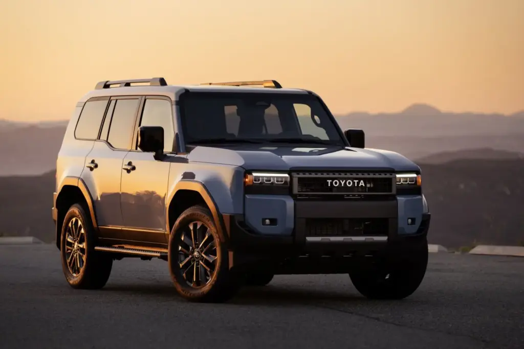 Toyota ha presentado en EE. UU. el Toyota Land Cruiser 2027 para el mercado norteamericano, una actualización que introduce mejoras concretas sobre la versión anterior. El precio de salida es de 57.880 dólares (unos 49.500 euros al cambio actual), y su llegada a los concesionarios del país presidido por Donald Trump está prevista para la primavera de 2026. Las novedades son moderadas pero relevantes. Entre los equipamientos disponibles por primera vez figura una toma de sdmisión elevada —también conocida como snorkel— y asientos calefactados y ventilados para las plazas exteriores de la segunda fila. Se suma un nuevo color de carrocería denominado Inked, una especie de negro pizarra que amplía la paleta disponible hasta siete opciones. El Toyota Land Cruiser 250 norteamericano frente al europeo En la actualidad, el Land Cruiser que se comercializa en EE. UU. es esencialmente el mismo modelo que el que se vende en Europa, pero difiere en aspectos fundamentales. El más llamativo es la mecánica: el modelo norteamericano incorpora en exclusiva el sistema híbrido i-Force Max, que combina un motor de gasolina V6 biturbo de 3,5 litros con un motor eléctrico integrado entre el propulsor y la transmisión automática de ocho velocidades. La potencia combinada es de 326 CV (240 kW), y el par máximo alcanza los 630 Nm (465 lb-ft). Toyota estima un consumo de 12,28 l/100 km (23 MPG combinados, según el ciclo EPA), que serían equivalentes a unos 10,2 l/100 km según el protocolo WLTP. El Land Cruiser europeo, en cambio, se ofrece actualmente con un diésel mild hybrid de 2.755 cm³ y 204 CV (150 kW) que homologa 10,2 litros de gasóleo cada 100 km en ciclo combinado WLTP. La tracción total permanente, con diferencial central y trasero bloqueables, es común a ambas versiones. Dos acabados y un equipamiento de serie que no deja de crecer El Toyota Land Cruiser 2027 se ofrece en EE. UU. en dos acabados. El denominado 1958 rinde homenaje al año de nacimiento del modelo con faros redondos de LED, tapicería de tela calefactada y la icónica parrilla con las letras «TOYOTA» en horizontal. El acabado Land Cruiser añade faros rectangulares, tapicería SofTex, asientos delanteros ventilados, cargador inalámbrico Qi, pantalla de 12,3 pulgadas y luces antiniebla LED Rigid multi-color. El paquete Premium, disponible sobre este segundo acabado, incorpora tapicería de cuero, ventilación también en los asientos traseros exteriores, sistema de audio JBL de 14 altavoces, espejo retrovisor digital y techo solar. La seguridad activa de serie incluye el paquete Toyota Safety Sense 3.0, con frenada automática de emergencia con detección de peatones, ciclistas y motociclistas, control de crucero adaptativo a todas las velocidades, asistente de mantenimiento de carril y reconocimiento de señales de tráfico. Incorpora además una función denominada Emergency Driving Stop, que monitoriza los datos del conductor y, si detecta falta de respuesta, detiene el vehículo de forma autónoma. La capacidad de remolque se sitúa en 2.722 kg (6.000 libras), y la garantía del sistema híbrido cubre ocho años o 160.000 km (100.000 millas), con la batería garantizada durante diez años o 240.000 km (150.000 millas). Las claves del Toyota Land Cruiser 2027 para EE. UU. El Toyota Land Cruiser 2027 americano incorpora una toma de aire elevada, asientos traseros ventilados y un nuevo color, Inked. Su precio de salida en EE. UU. es de 57.880 dólares. El motor es el híbrido 3.5 V6 biturbo, con 326 CV (240 kW) y 630 Nm, exclusivo del mercado norteamericano. Se ofrecen dos acabados (1958 y Land Cruiser) y un paquete Premium con audio JBL de 14 altavoces y techo solar. La garantía de la batería híbrida cubre diez años o 240.000 km.