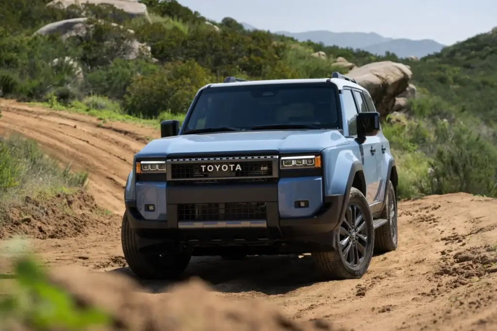 Toyota ha presentado en EE. UU. el Toyota Land Cruiser 2027 para el mercado norteamericano, una actualización que introduce mejoras concretas sobre la versión anterior. El precio de salida es de 57.880 dólares (unos 49.500 euros al cambio actual), y su llegada a los concesionarios del país presidido por Donald Trump está prevista para la primavera de 2026. Las novedades son moderadas pero relevantes. Entre los equipamientos disponibles por primera vez figura una toma de sdmisión elevada —también conocida como snorkel— y asientos calefactados y ventilados para las plazas exteriores de la segunda fila. Se suma un nuevo color de carrocería denominado Inked, una especie de negro pizarra que amplía la paleta disponible hasta siete opciones. El Toyota Land Cruiser 250 norteamericano frente al europeo En la actualidad, el Land Cruiser que se comercializa en EE. UU. es esencialmente el mismo modelo que el que se vende en Europa, pero difiere en aspectos fundamentales. El más llamativo es la mecánica: el modelo norteamericano incorpora en exclusiva el sistema híbrido i-Force Max, que combina un motor de gasolina V6 biturbo de 3,5 litros con un motor eléctrico integrado entre el propulsor y la transmisión automática de ocho velocidades. La potencia combinada es de 326 CV (240 kW), y el par máximo alcanza los 630 Nm (465 lb-ft). Toyota estima un consumo de 12,28 l/100 km (23 MPG combinados, según el ciclo EPA), que serían equivalentes a unos 10,2 l/100 km según el protocolo WLTP. El Land Cruiser europeo, en cambio, se ofrece actualmente con un diésel mild hybrid de 2.755 cm³ y 204 CV (150 kW) que homologa 10,2 litros de gasóleo cada 100 km en ciclo combinado WLTP. La tracción total permanente, con diferencial central y trasero bloqueables, es común a ambas versiones. Dos acabados y un equipamiento de serie que no deja de crecer El Toyota Land Cruiser 2027 se ofrece en EE. UU. en dos acabados. El denominado 1958 rinde homenaje al año de nacimiento del modelo con faros redondos de LED, tapicería de tela calefactada y la icónica parrilla con las letras «TOYOTA» en horizontal. El acabado Land Cruiser añade faros rectangulares, tapicería SofTex, asientos delanteros ventilados, cargador inalámbrico Qi, pantalla de 12,3 pulgadas y luces antiniebla LED Rigid multi-color. El paquete Premium, disponible sobre este segundo acabado, incorpora tapicería de cuero, ventilación también en los asientos traseros exteriores, sistema de audio JBL de 14 altavoces, espejo retrovisor digital y techo solar. La seguridad activa de serie incluye el paquete Toyota Safety Sense 3.0, con frenada automática de emergencia con detección de peatones, ciclistas y motociclistas, control de crucero adaptativo a todas las velocidades, asistente de mantenimiento de carril y reconocimiento de señales de tráfico. Incorpora además una función denominada Emergency Driving Stop, que monitoriza los datos del conductor y, si detecta falta de respuesta, detiene el vehículo de forma autónoma. La capacidad de remolque se sitúa en 2.722 kg (6.000 libras), y la garantía del sistema híbrido cubre ocho años o 160.000 km (100.000 millas), con la batería garantizada durante diez años o 240.000 km (150.000 millas). Las claves del Toyota Land Cruiser 2027 para EE. UU. El Toyota Land Cruiser 2027 americano incorpora una toma de aire elevada, asientos traseros ventilados y un nuevo color, Inked. Su precio de salida en EE. UU. es de 57.880 dólares. El motor es el híbrido 3.5 V6 biturbo, con 326 CV (240 kW) y 630 Nm, exclusivo del mercado norteamericano. Se ofrecen dos acabados (1958 y Land Cruiser) y un paquete Premium con audio JBL de 14 altavoces y techo solar. La garantía de la batería híbrida cubre diez años o 240.000 km.